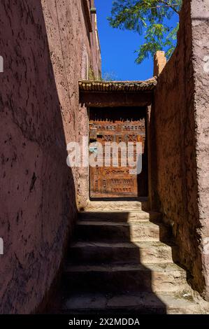 Une vieille porte en bois ornée au cœur du Maroc, avec des sculptures élaborées, encadrées par des murs roses altérés et des marches en pierre sous le ciel bleu clair. Banque D'Images