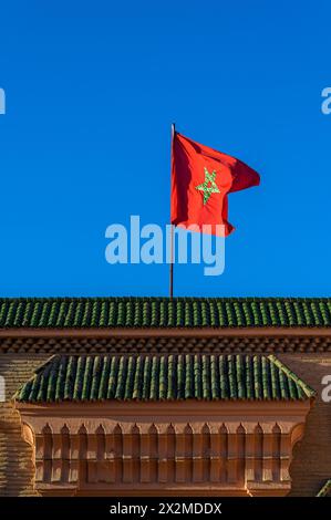 Le vibrant drapeau marocain flotte fièrement dans un ciel sans nuages au-dessus d'un bâtiment à l'architecture marocaine traditionnelle. Banque D'Images