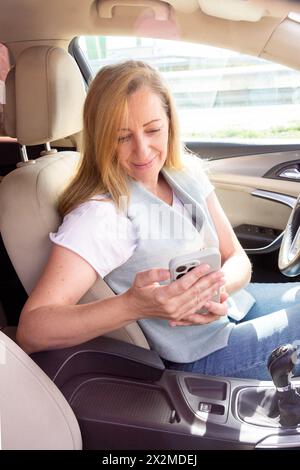 Une femme d'âge mûr souriante s'assoit sur le siège passager d'une voiture, en train d'interagir avec son smartphone Banque D'Images
