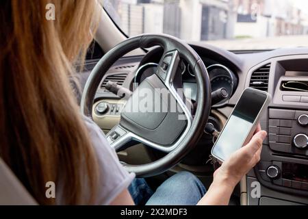 Une conductrice est photographiée tenant un smartphone, sur le point de l’utiliser assise dans une voiture garée, suggérant un moment de navigation ou de communication Banque D'Images