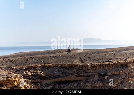 Vue de côté d'une femme active randonne à travers le paysage aride du sud de Fuerteventura, avec l'océan Atlantique scintillant et les montagnes lointaines en t Banque D'Images