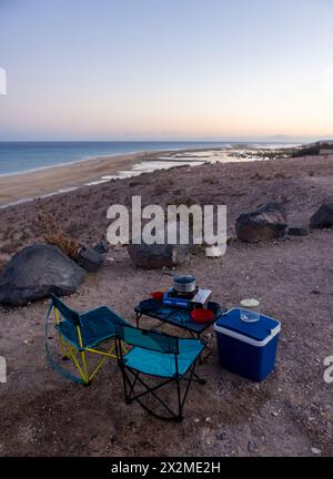 Une scène de camping tranquille avec une chaise, une cuisinière et une glacière donnant sur la plage au crépuscule sur l'île de Fuerteventura, en Espagne. Banque D'Images