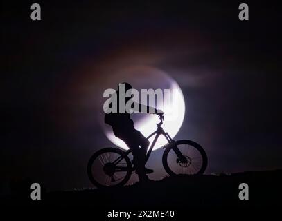 Silhouette d'un cycliste avec un VTT contre un ciel plein clair de lune, véhiculant l'aventure et l'esprit des balades nocturnes d'été Banque D'Images