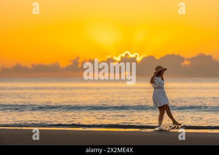 Vue latérale d'une femme profite d'une promenade paisible sur une plage de sable avec un coucher de soleil vibrant en arrière-plan. Banque D'Images