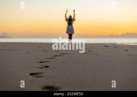 Silhouette méconnaissable d'une femme aux bras levés, debout sur une plage calme à l'aube, exprimant la liberté et la détente. Banque D'Images