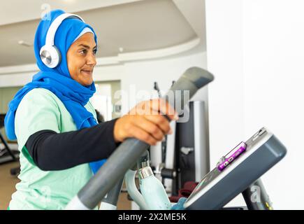 Une femme arabe d'âge moyen avec un hijab bleu utilisant un tapis roulant à la salle de gym tout en portant des écouteurs, engagée dans sa routine d'entraînement Banque D'Images