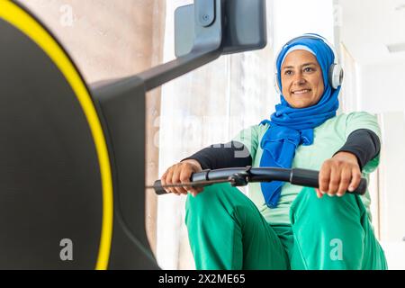 Une femme arabe dans un hijab et un casque sourit tout en s'entraînant sur des équipements de gymnastique, illustrant la diversité dans la forme physique Banque D'Images