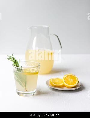 Un pichet plein de limonade rafraîchissante à côté d'un verre rempli garni de romarin, avec des citrons coupés en deux sur une soucoupe sur un fond blanc Banque D'Images