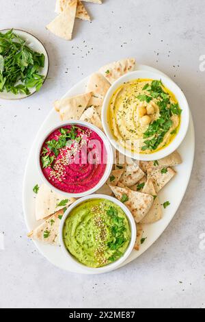 Vue de dessus trois bols de houmous maison coloré - classique, betterave et avocat - servis avec du pain pita et garnis d'herbes. Banque D'Images