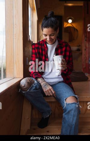 Une femme souriante avec des tatouages est assise près d'une fenêtre, tenant une tasse de café dans un intérieur confortable et en bois, exsudant une ambiance détendue Banque D'Images