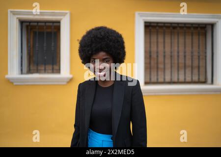 Une jeune femme avec un sourire radieux et une coiffure afro posant avec confiance devant un mur jaune, dégageant une ambiance de style de vie urbain dynamique Banque D'Images