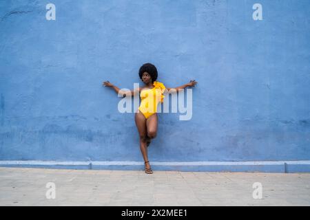 Une femme afro confiante pose dans un monobloc jaune vif sur un fond bleu texturé, dégageant style audacieux et énergie Banque D'Images