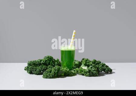 Un jus de chou frisé vert vif servi dans un verre transparent avec une paille rayée jaune, accompagné de feuilles de chou frisé frais sur un fond neutre Banque D'Images