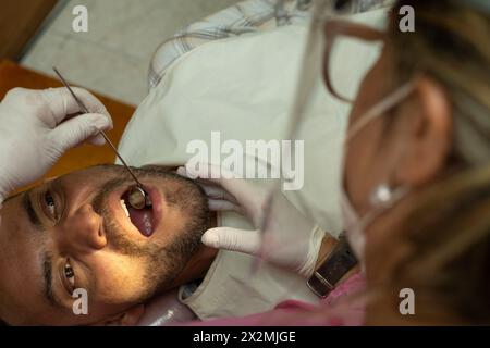 Un homme adulte latino-américain (42) est examiné par une dentiste à l'aide d'un miroir dentaire. vue zénithale. Concept de santé et odontologie. Banque D'Images