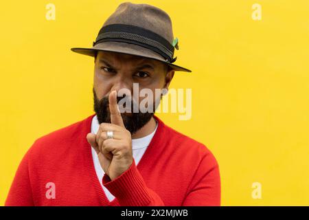 Adulte Latino mâle (39 ans) avec barbe, faisant un geste silencieux avec son doigt droit devant sa bouche. Il porte un chapeau, un pull rouge et un fla blanc Banque D'Images