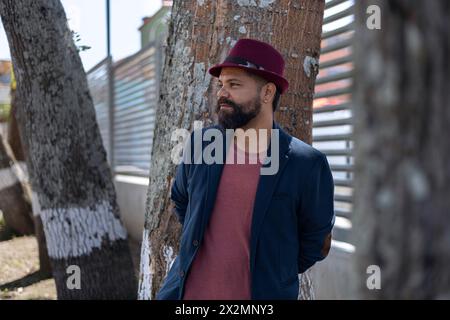 Plan moyen long de l'homme bohème latino-américain avec barbe, chapeau et veste debout à côté de quelques arbres dans la ville. Concept de vacances et portrait Banque D'Images