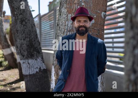 Plan moyen long de l'homme bohème latino-américain avec barbe, chapeau et veste debout à côté de quelques arbres dans la ville. Concept de vacances et portrait Banque D'Images