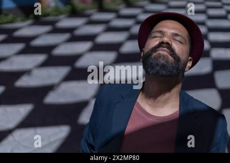 Portrait d'homme bohème latino-américain avec les yeux fermés, barbe, chapeau et veste. Fond d'ombres avec des formes de losange. Concept de vraies personnes. Banque D'Images