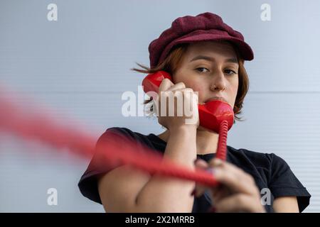 Plan court moyen d'une jeune femme latino-américaine (22 ans) avec casquette et cheveux roux écoutant la conversation sur le combiné rouge rétro. Prise de vue à angle bas. Vintage tec Banque D'Images