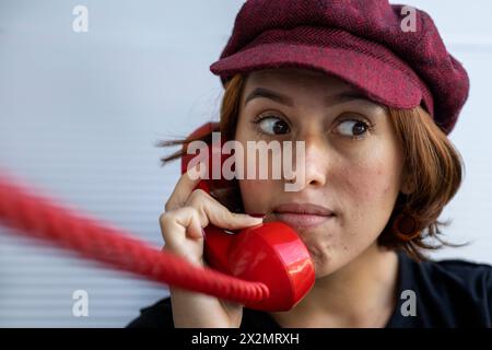 Plan court moyen d'une jeune fille latino-américaine (22 ans) avec une casquette et les cheveux roux écoutant un potins sur son combiné rouge rétro et faisant un geste surpris Banque D'Images