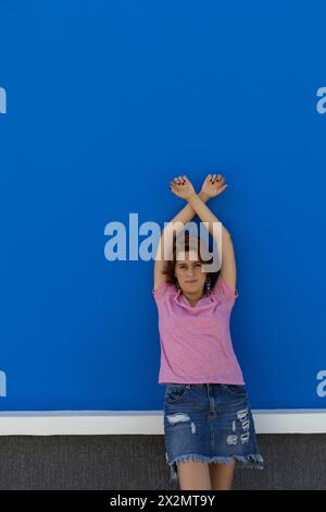 Plan moyen long d'une jeune femme (22) rousse latino-américaine regardant et faisant face à la caméra avec ses bras tendus vers le haut. Fond de mur bleu. Portrait c Banque D'Images