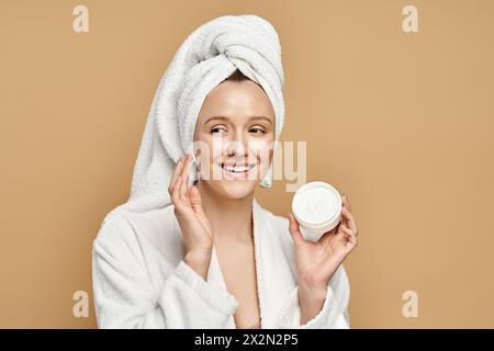 Une femme avec une serviette sur la tête tient un pot de crème, mettant en valeur sa routine beauté dans un cadre serein. Banque D'Images