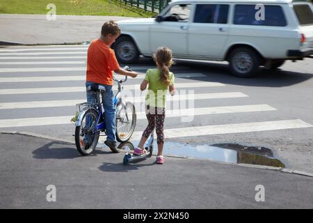 Garçon avec vélo et sa jeune sœur avec scooter stand avant la traversée zèbre avec voiture en mouvement Banque D'Images