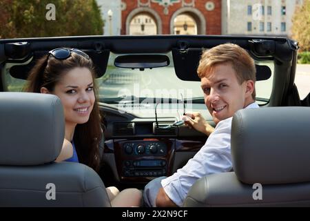 Jeune couple, regardant en arrière, est assis dans un cabriolet contre le bâtiment avec deux arches, la fille est sur un siège conducteur. Banque D'Images