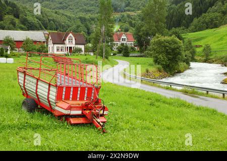 Vieux telega rouge près de la route du village parmi les arbres près de la rivière sous la montagne Banque D'Images