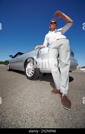 Jeune homme dans des lunettes de soleil sombres se tient penché son coude sur le coffre d'une voiture argentée à toit ouvert et pairs loin Banque D'Images