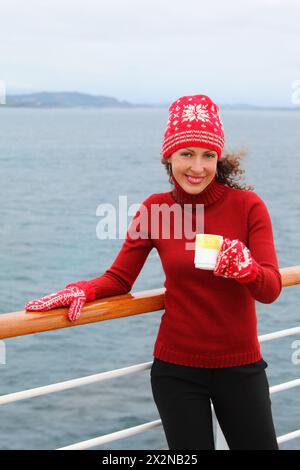 Femme souriante portant un chapeau rouge et des mitaines tient une tasse de thé chaud au pont du navire Banque D'Images