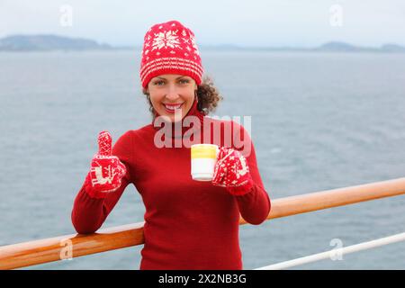 Femme portant un chapeau rouge et des mitaines tient une tasse de thé chaud et les pouces vers le haut au pont du navire Banque D'Images