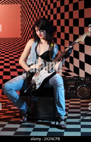 Femmes avec guitare électrique est assis sur l'amplificateur dans le studio avec fond damier et lumière rouge. Banque D'Images