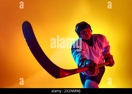 Joueur de hockey sur glace concentré posant avec un bâton montrant son prêt à gagner dans la lumière néon sur fond dégradé jaune. Banque D'Images