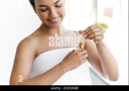 Gros plan d'une jolie jeune femme hispanique ou brésilienne, enveloppée dans une serviette blanche, appliquant un sérum hydratant sur ses mains après la douche, souriante. Prendre soin d'un corps et des mains Banque D'Images