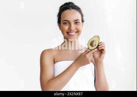 Charmante jeune femme brésilienne ou hispanique bien entretenue enveloppée dans une serviette se tient sur un fond blanc, tient un avocat dans ses mains, regarde et sourit à la caméra Banque D'Images