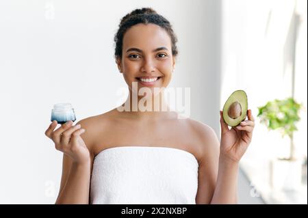 Belle charmante jeune femme brésilienne ou hispanique bien entretenue enveloppée dans une serviette se tient sur fond blanc, tient un avocat et une crème hydratante dans ses mains, sourire à la caméra. Concept de soin de la peau Banque D'Images