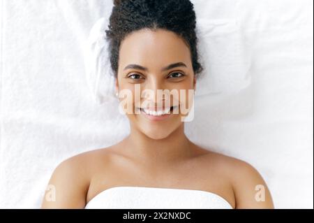 Vue de dessus d'une belle jeune femme brésilienne ou hispanique bien soignée enveloppée dans une serviette, couchée dans un salon de beauté avant un massage ou une procédure de soins de la peau du visage, regardant la caméra, souriant Banque D'Images