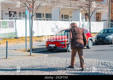 Un vieil homme avec un bâton de marche traverse une rue, photographié par derrière Banque D'Images