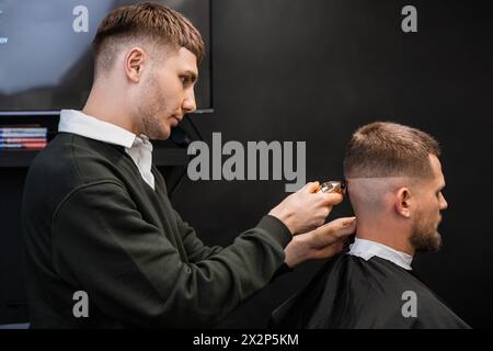 Le jeune barbier coupe les cheveux de l'homme en utilisant la tondeuse dans le salon de coiffure. Banque D'Images