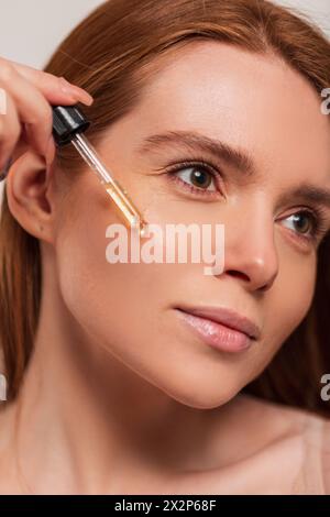 Belle jeune jeune femme rousse fraîche avec peau faciale claire gouttes d'huile d'Un compte-gouttes sur sa peau, en gros plan. Beauté et soins de la peau Banque D'Images