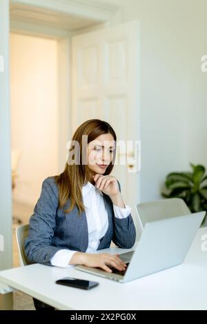 Femme professionnelle concentrée travaille avec diligence sur son ordinateur portable, son espace de travail baigné de lumière naturelle. Banque D'Images