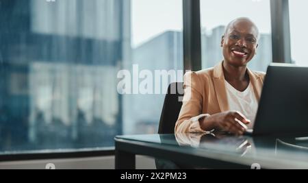 Femme d'affaires heureuse et prospère utilisant un ordinateur portable dans son bureau. Elle est assise à une table, souriant et regardant la caméra avec confiance. Sa baguette professionnelle Banque D'Images