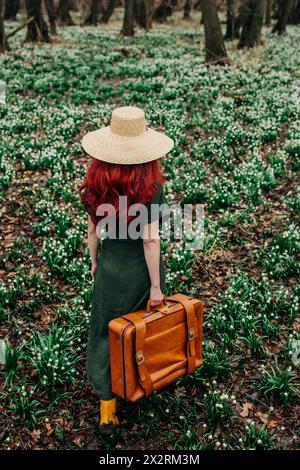 Femme marchant avec une valise vintage à travers les fleurs de Lily-of-the-Valley dans la forêt Banque D'Images