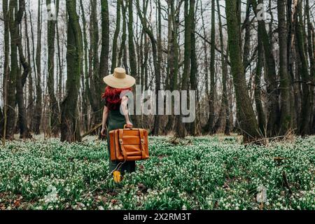 Femme marchant avec une valise vintage à travers des fleurs dans la forêt Banque D'Images