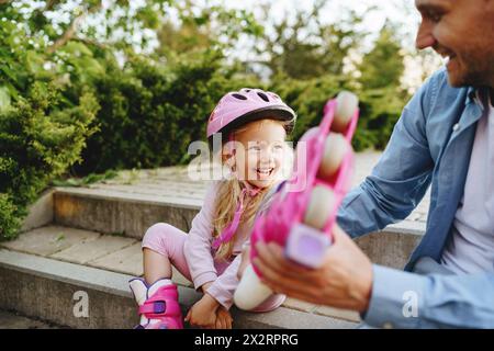 Jeune fille joyeuse aidée par son père à porter des patins à roulettes assis sur des marches Banque D'Images