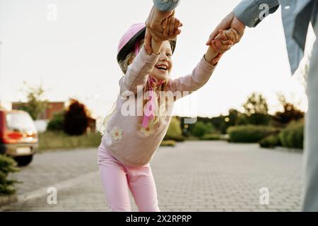 Jeune fille joyeuse tenant la main de son père et apprenant à patiner à roulettes sur un sentier pédestre Banque D'Images