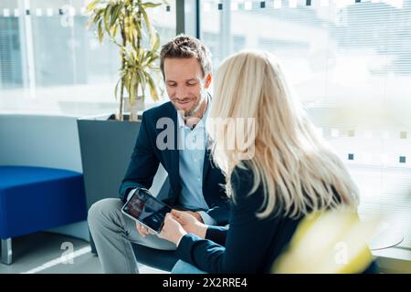 Homme d'affaires souriant assis avec femme d'affaires tenant la tablette PC dans le bureau Banque D'Images