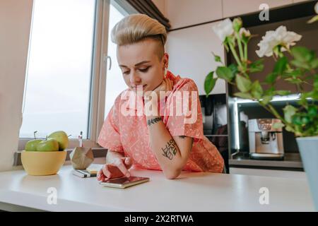 Femme appuyée sur le coude et défilant téléphone intelligent dans la cuisine à la maison Banque D'Images