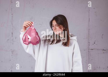 Fille tenant un ballon rose en forme de coeur dégonflé devant un fond de ciment Banque D'Images